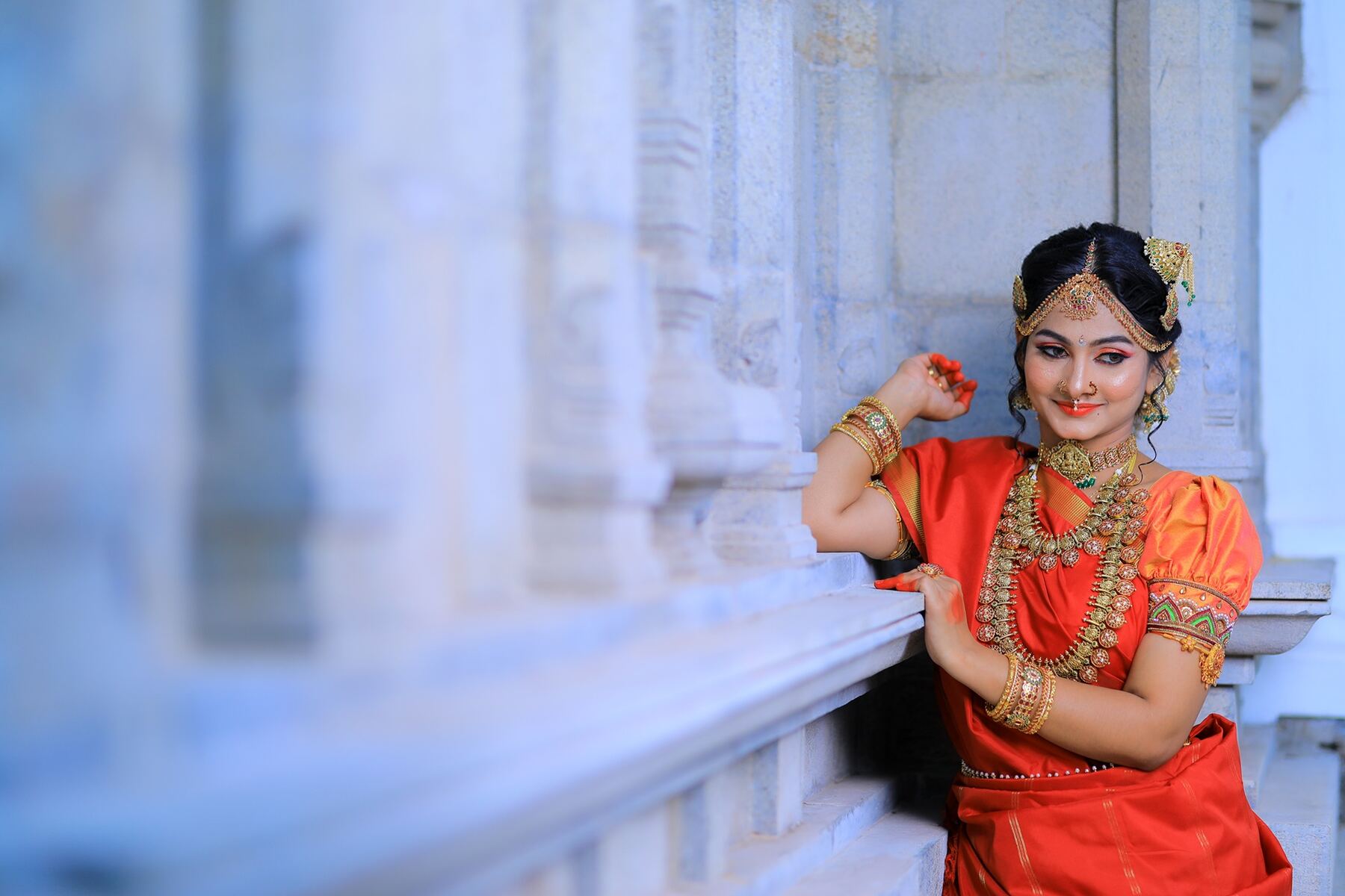 Bride in orange silk saree seated in a temple corridor after bridal makeup